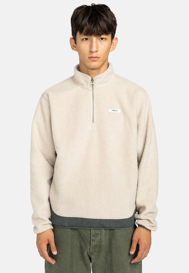 Polarowy pulower w jasnym beżu z półzamkiem, wysokim kołnierzem i kontrastowym, ciemnozielonym wykończeniem. Posiada małe logo na klatce piersiowej.