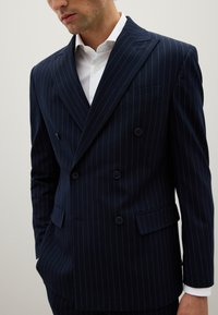 Chaqueta de traje azul marino de doble botonadura con rayas blancas, diseño estructurado, solapas de picos y dos bolsillos frontales. Textura de tela suave.