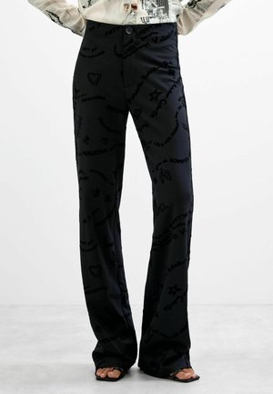 Pantalon noir avec texte et symboles imprimés subtilement, porté avec des sandales noires à bouts ouverts et un chemisier léger à motifs.