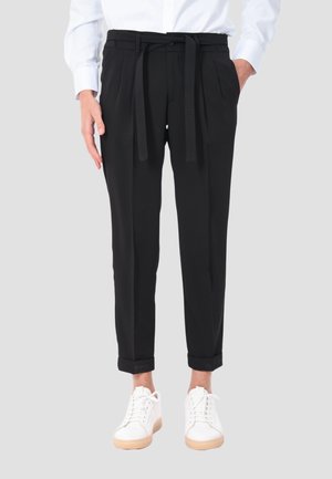 Persona che indossa pantaloni neri a vita alta con cintura di stoffa, orli risvoltati, camicia bianca infilata e sneakers bianche con suola marrone chiaro.