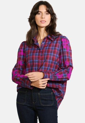 Ruitjesoverhemd in rood en blauw met lange mouwen, voorzien van levendige roze bloemenborduursels op de schouders. Donkere jeans eronder gedragen.