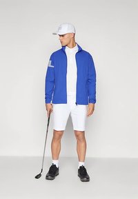 Άνδρας με λευκό καπέλο, μπλε μπουφάν, λευκό πόλο και σορτς, κρατώντας ένα γκολφ ρόπαλο, στέκεται σε απλό φωτεινό φόντο.