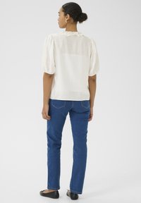 Chemisier ample en ivoire avec des manches courtes bouffantes et un col à volants, associé à un jean droit en denim bleu et des ballerines noires.