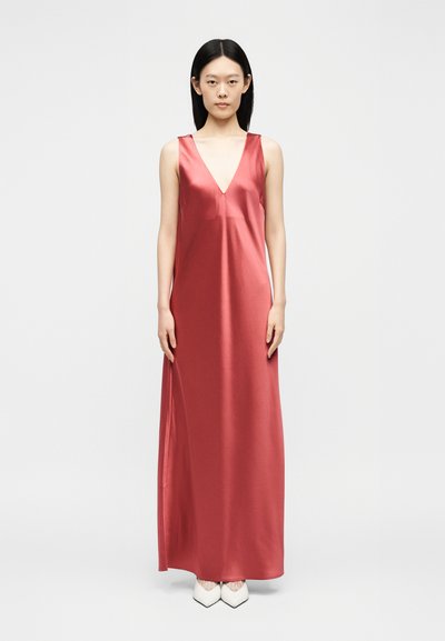 Γυναίκα με ίσια μαύρα μαλλιά που φοράει αμάνικο μακρύ κόκκινο σατέν φόρεμα με βαθύ V ντεκολτέ και λευκά μυτερά παπούτσια, στέκεται σε λευκό φόντο.