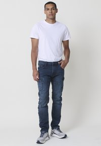 Camiseta ligera de algodón blanca combinada con pantalones cargo de mezclilla azules que cuentan con múltiples bolsillos y un corte estrecho. Zapatillas de deporte grises completan el look.