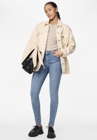 Beige oversized denim jack met een gestreept shirt, gecombineerd met lichtblauwe skinny jeans en zwarte chunky schoenen. Zwarte tas met beige sjaal.