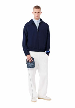 Homme portant une veste zippée bleu marine, pantalon blanc, baskets blanches, tenant une petite pochette à motifs avec un logo, sur fond blanc.