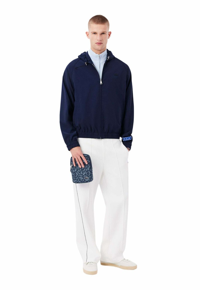 Homme portant une veste zippée bleu marine, pantalon blanc, baskets blanches, tenant une petite pochette à motifs avec un logo, sur fond blanc.