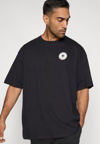 Fekete pamut póló rövid ujjal, kerek nyakkivágással és egy kör alakú logópárnával a bal mellrészen. Laza szabású, sima textúrájú.