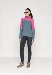 Veste légère en teal et rose, à fermeture éclair avec un logo ; associée à des leggings noirs et des chaussures de randonnée marron. Des lunettes de soleil complètent le look.