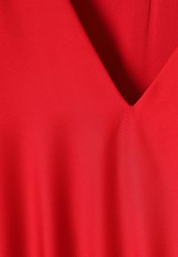 Jasne červené tkaniny s hlbokým výstrihom do V, hladkou štruktúrou a jemnými záhybmi zvýrazňujúcimi tvar odevu.