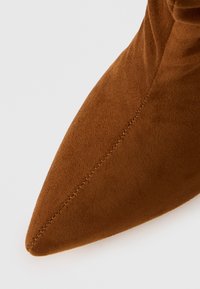Bottine en daim marron avec un bout pointu, présentant une texture lisse et des coutures discrètes le long du bord.