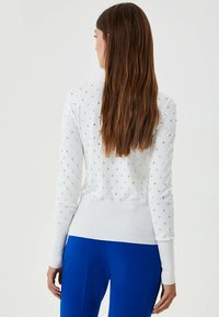 Weißer Strickpullover mit langen Ärmeln, der ein strukturiertes Muster aus kleinen silbernen Verzierungen und einen enganliegenden, gerippten Bundsaum aufweist.