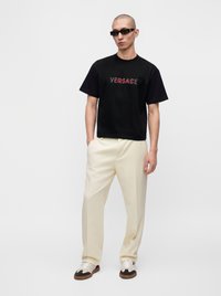 Φαλακρός άνδρας που φοράει μαύρο μπλουζάκι Versace, κρεμ παντελόνι, γυαλιά ηλίου και λευκά αθλητικά παπούτσια στέκεται μπροστά σε απλό λευκό φόντο.