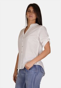 Camicia bianca di lino con colletto alla mandarino, apertura con bottoni e maniche arrotolate. Indossata con jeans in denim, con una vestibilità comoda e una consistenza morbida.