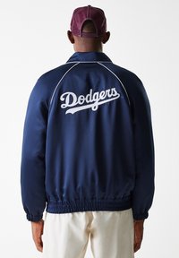 Blue satin baseball jacket with white "Dodgers" logo, elastic cuffs, and waistband. Features contrasting white piping along shoulder seam.