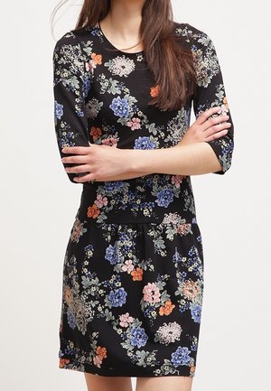 Vrouw in een zwarte jurk met blauwe, roze, oranje en witte bloemmotieven, armen gekruist en driekwartmouwen.