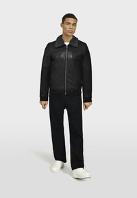 Chaqueta bomber de cuero negra con collar de piel gris, cierre de cremallera, puños acanalados, combinada con pantalones oscuros y zapatillas blancas. El modelo está de pie.