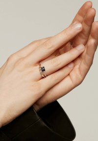 Anillo de oro con una piedra negra central flanqueada por pequeñas gemas transparentes, colocado en una mano. La mano está posicionada suavemente contra otra mano.