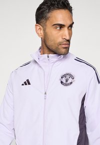 Jasnofioletowa kurtka na zamek z czarnymi akcentami i logo Manchester United na piersi. Posiada kieszenie na zamek i paski na ramionach.