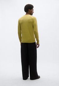 Κίτρινο πλεκτό πουλόβερ με στρογγυλή λαιμόκοψη, συνδυασμένο με φαρδιά μαύρη παντελόνα. Το πουλόβερ έχει λεία υφή και λιτές λεπτομέρειες σχεδίασης.