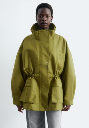 Personne portant une veste oversize vert olive avec un col haut et de grandes poches avant, associée à un pantalon gris clair sur un fond uni.