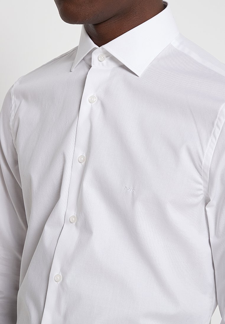 Camisa blanca de manga larga con cierre de botones, que presenta un cuello clásico y un logotipo sutilmente bordado en el pecho. Textura de tela suave.