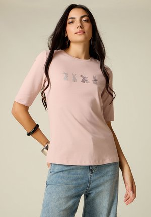Kobieta o długich ciemnych włosach, ubrana w jasnoróżowy t-shirt z pięcioma małymi nadrukami królików na wysokości piersi oraz niebieskie dżinsy, stojąca z ręką na biodrze.