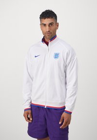 Biela bunda na zips, s modrou logom Nike a anglickým erbom. Rybkovaný spodný lem s červenými a modrými akcentami. Nosí sa cez marónovú košeľu a fialové šortky.