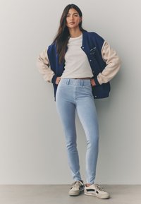 Modro-bež univerzitetna jakna z mehki rokavi, bela majica, svetlo modre ozke kavbojke in bež superge s črnimi poudarki.