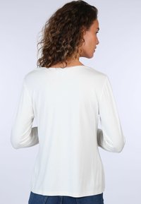 Leichtes, cremefarbenes Langarmshirt mit Rundhalsausschnitt und Spitzenverzierung. Hat einen lockeren Sitz und eine glatte Textur. Die Rückansicht zeigt minimale Details.