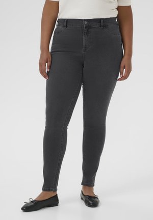 Vrouw draagt donkergrijze skinny jeans en zwarte ballerina's tegen een effen lichte achtergrond.