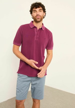 Mann mit lockigem Haar, der ein violettes Poloshirt und hellblaue Shorts trägt, steht vor einer schlichten Wand und gestikuliert mit den Händen.