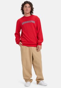 Červená mikina s logom "QUIKSILVER"; okrúhly výstrih, dlhé rukávy. V kombinácii s béžovými, voľne strihanými nohavicami a bielymi topánkami. Hladká textúra.