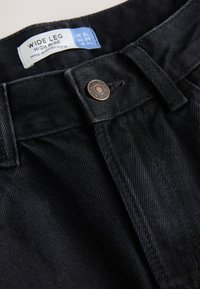 Črne široke kavbojke z visokim pasom iz jeansa, z bronasto gumbom in etiketo z znamko ter podatki o velikosti. Tekstura je videti trpežna in gladka.