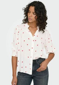 Camisa blanca de botones con mangas cortas acampanadas y estampado de corazones rojos. Tejido ligero con un ajuste relajado, combinada con jeans de mezclilla oscura.