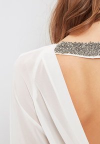 Espalda de una mujer con una blusa blanca transparente que presenta una espalda descubierta y un escote adornado con pequeñas cuentas plateadas.