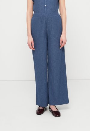 Μπλε παντελόνια φαρδιάς γραμμής με μοτίβο και υφή υφάσματος, με ψηλή μέση και κούμπωμα με κουμπιά μπροστά. Συνδυασμένα με μπορντό παπούτσια.