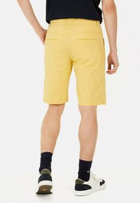 Gule shorts med flad front og baglommer, med bæltestropper i taljen. Modellen har hvide sneakers og marineblå sokker på.