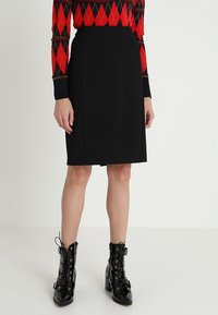 Femme portant une jupe crayon noire longueur genou, un pull en argyle rouge et noir, et des bottines à lacets en cuir verni noir.
