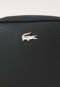 Crna teksturirana kožna torba s malim srebrnim logotipom krokodila na prednjoj strani. Glatka površina s strukturiranim oblikom.