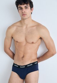 Marineblaue Hugo Slips mit einem schwarzen Bund, der den Markennamen in Weiß zeigt. Der Stoff wirkt glatt und passt sich dem Körper an.