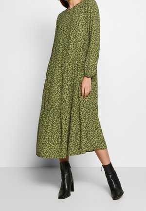 Femme portant une robe midi verte à manches longues avec un petit motif abstrait, associée à des bottines noires à talons carrés.