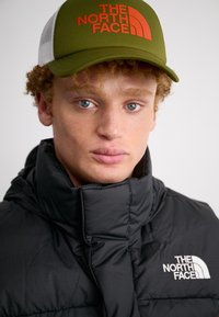 Doudoune noire avec col haut, arborant un logo blanc. Casquette de baseball verte avec un logo orange et un dos en maille. Yeux bleus et cheveux bouclés visibles.