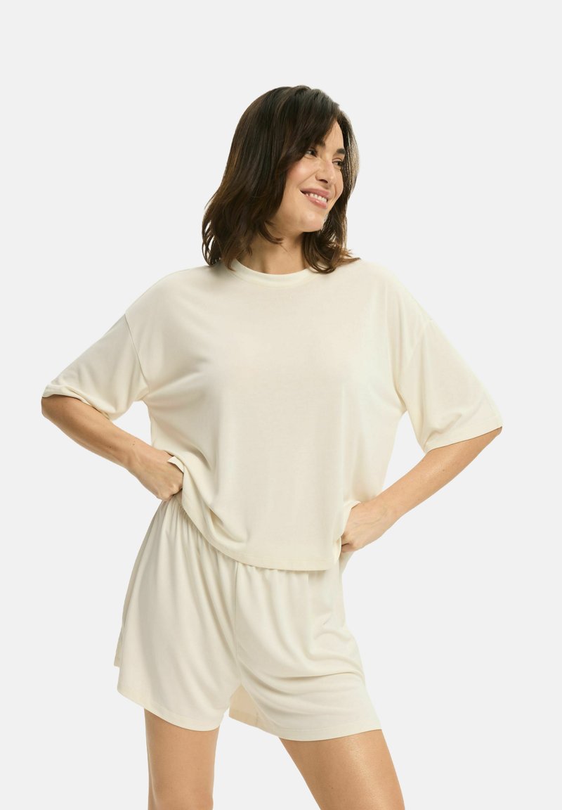 Kremowa bluzka z krótkim rękawem o luźnym kroju i okrągłym dekoltem, w komplecie z pasującymi szortami, wykonana z miękkiego, lekkiego materiału. Gładka tekstura.