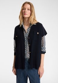 Marineblauer gestrickter Schlaf mit kurzen Ärmeln, zwei Fronttaschen und goldenen Knöpfen, über einer gemusterten blau-weißen Bluse getragen, kombiniert mit Jeanshosen.