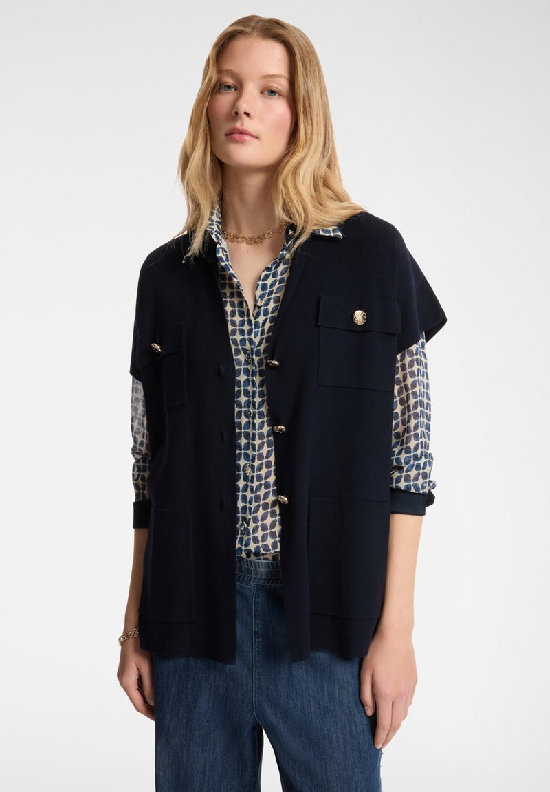 Marineblauer gestrickter Schlaf mit kurzen Ärmeln, zwei Fronttaschen und goldenen Knöpfen, über einer gemusterten blau-weißen Bluse getragen, kombiniert mit Jeanshosen.
