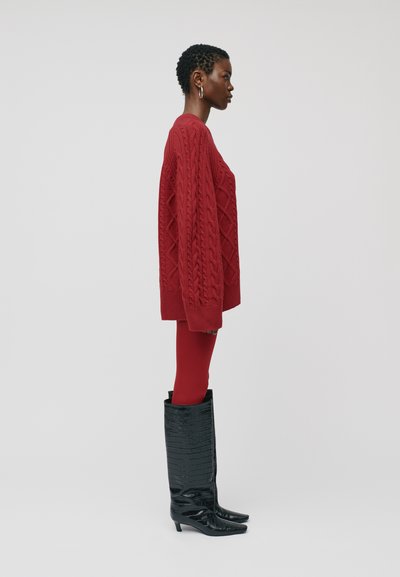 Moteris raudoname megztoje megztinyje ir legginguose, su juodais tekstūruotais iki kelių batais, stovinti šone prieš paprastą foną.