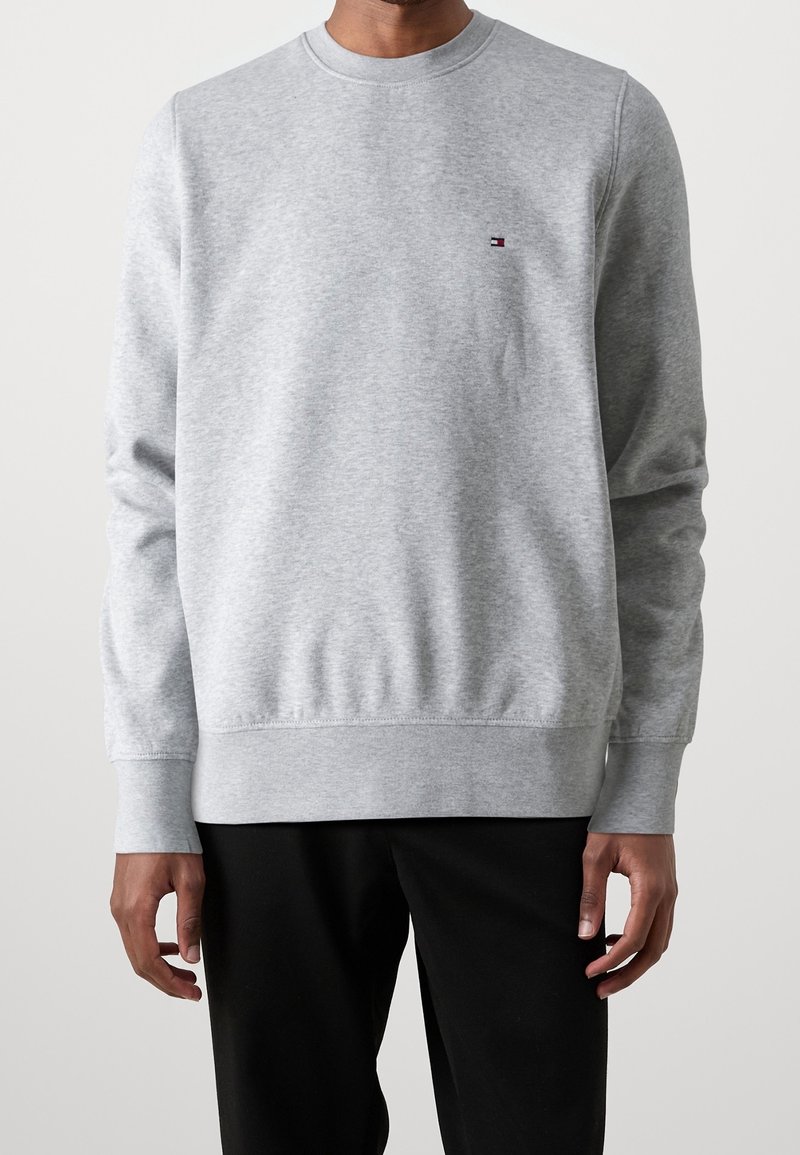 Homme portant un sweat-shirt gris clair à col ras du cou avec un petit logo rouge, blanc et bleu sur le côté gauche de la poitrine, associé à un pantalon noir.