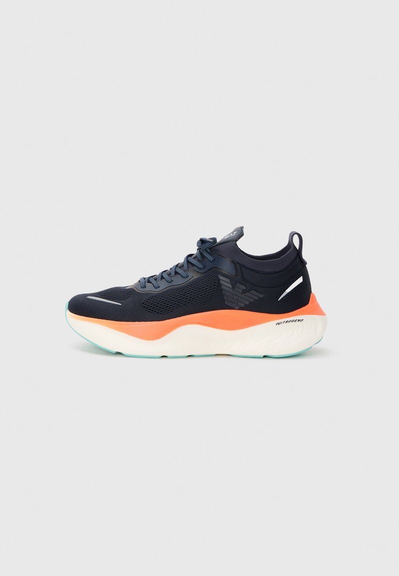 Baskets sportives noires avec semelle intermédiaire orange, semelle blanche, design à lacets et logo ailé sur le côté sur fond uni.